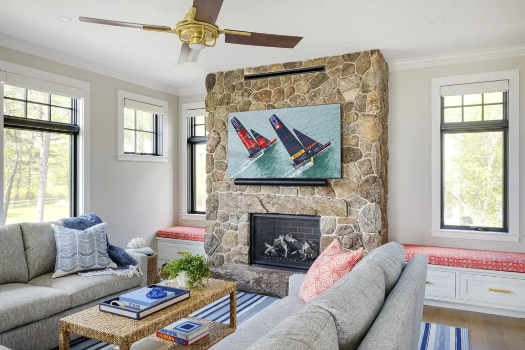 The living room introduces coral-colored fabric to the traditional blue and white color scheme. It centers on a fieldstone fireplace, in front of which are two custom sofas and a rattan-peel coffee table from Society Social.